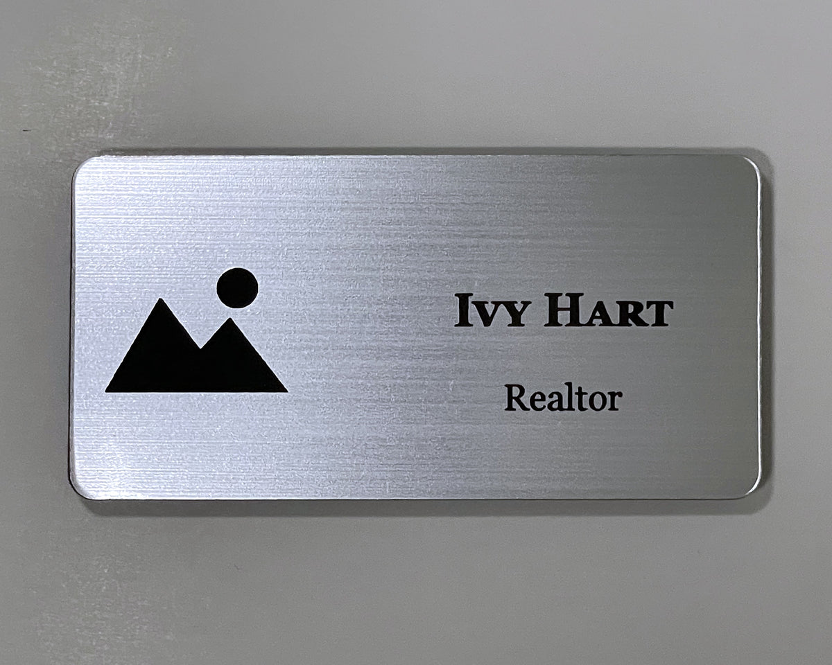 A brushed silver engraved realtor name badge with a black mountain and sun logo on the left and IVY HART, Realtor, on the right.
