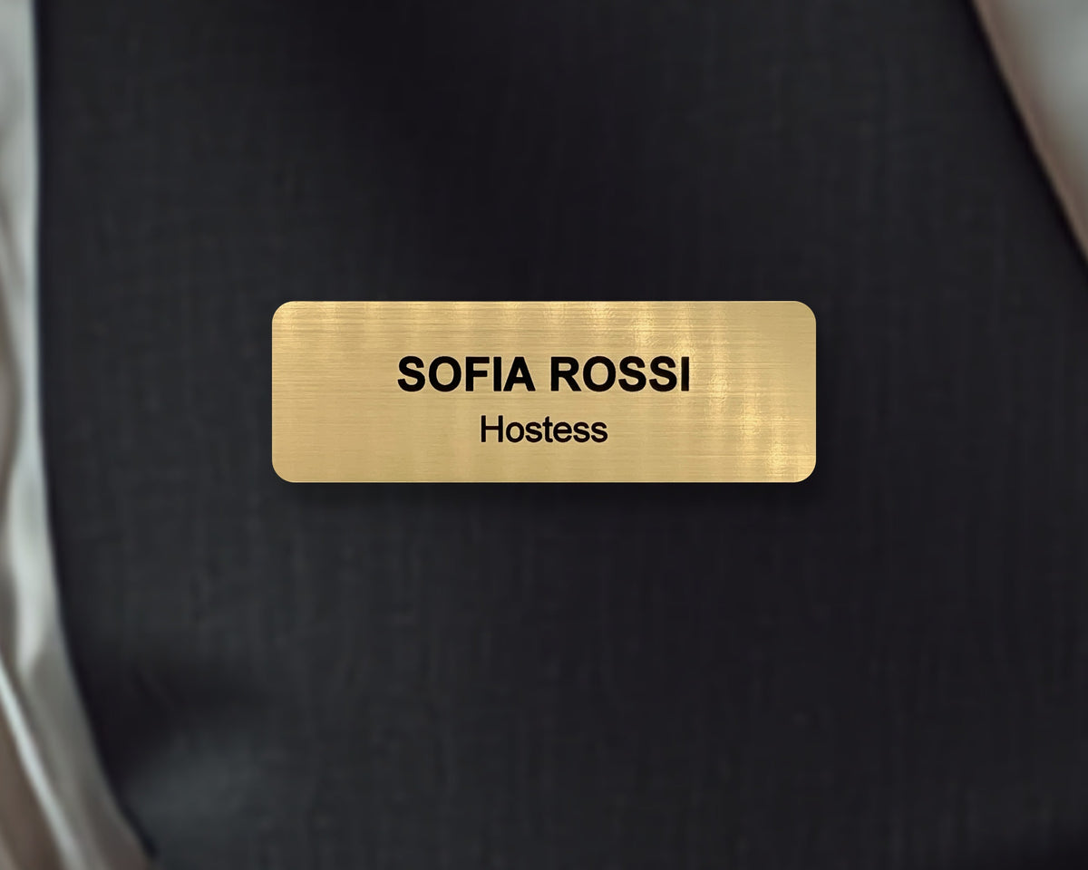 A rectangular gold-toned name badge with a brushed metal-look finish. The text SOFIA ROSSI is engraved in black capital letters, with the word Hostess below in smaller black letters. The badge is centered against a dark background.