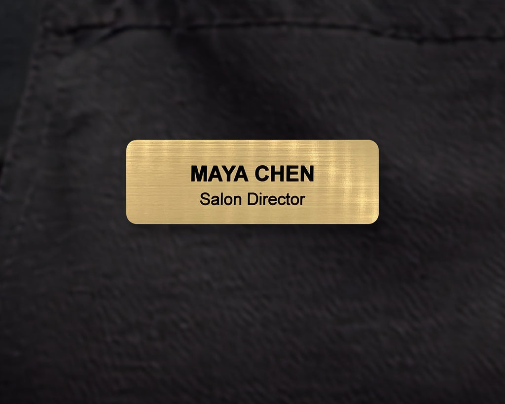 A rectangular gold-toned name badge with a brushed metal-look finish. The text reads MAYA CHEN in bold black letters, with Salon Director engraved below.