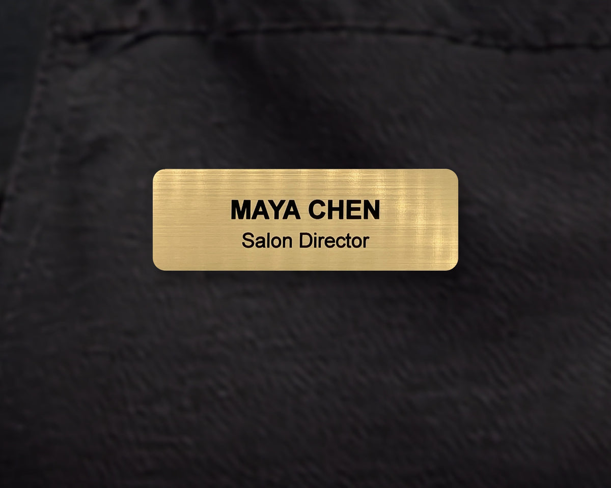 A rectangular gold-toned name badge with a brushed metal-look finish. The text reads MAYA CHEN in bold black letters, with Salon Director engraved below.