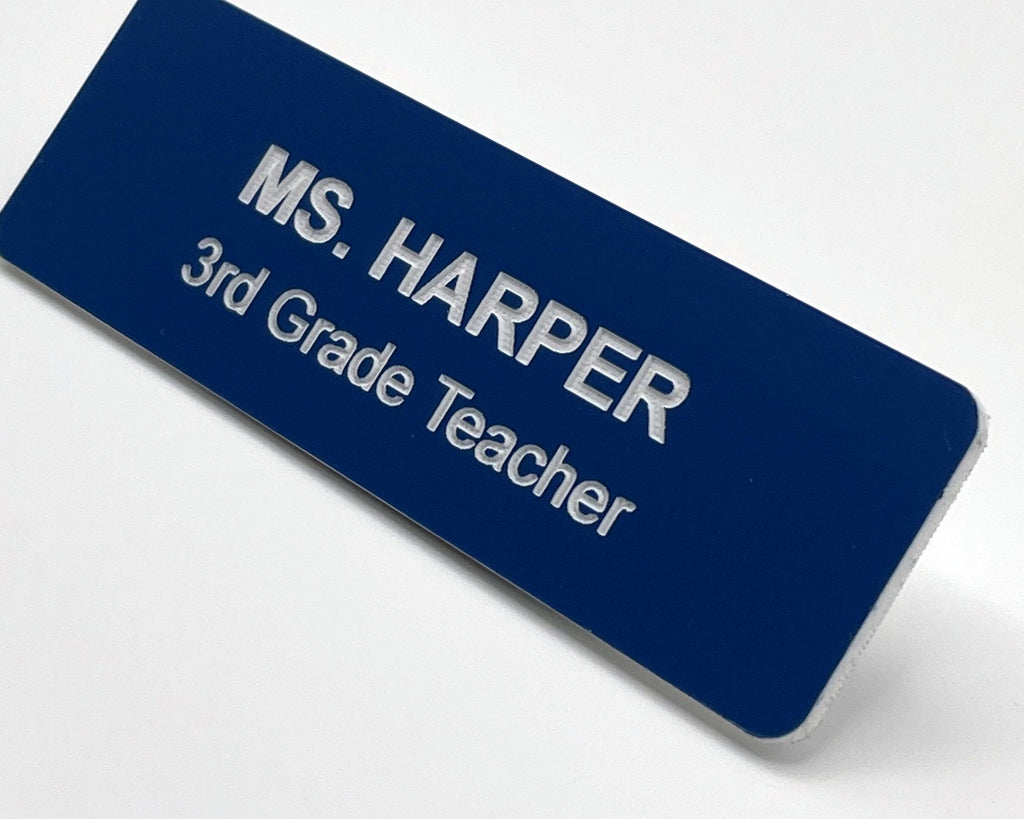 A rectangular dark blue name badge with white engraved text that reads MS. HARPER 3rd Grade Teacher. The badge has a clean, professional design with a smooth finish and rounded corners, clearly displaying the name and job title.
