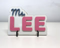 Teacher desk name sign reading Ms. LEE: big pink raised letters under a slate blue script Ms., on a white finish base that stands on two small feet.