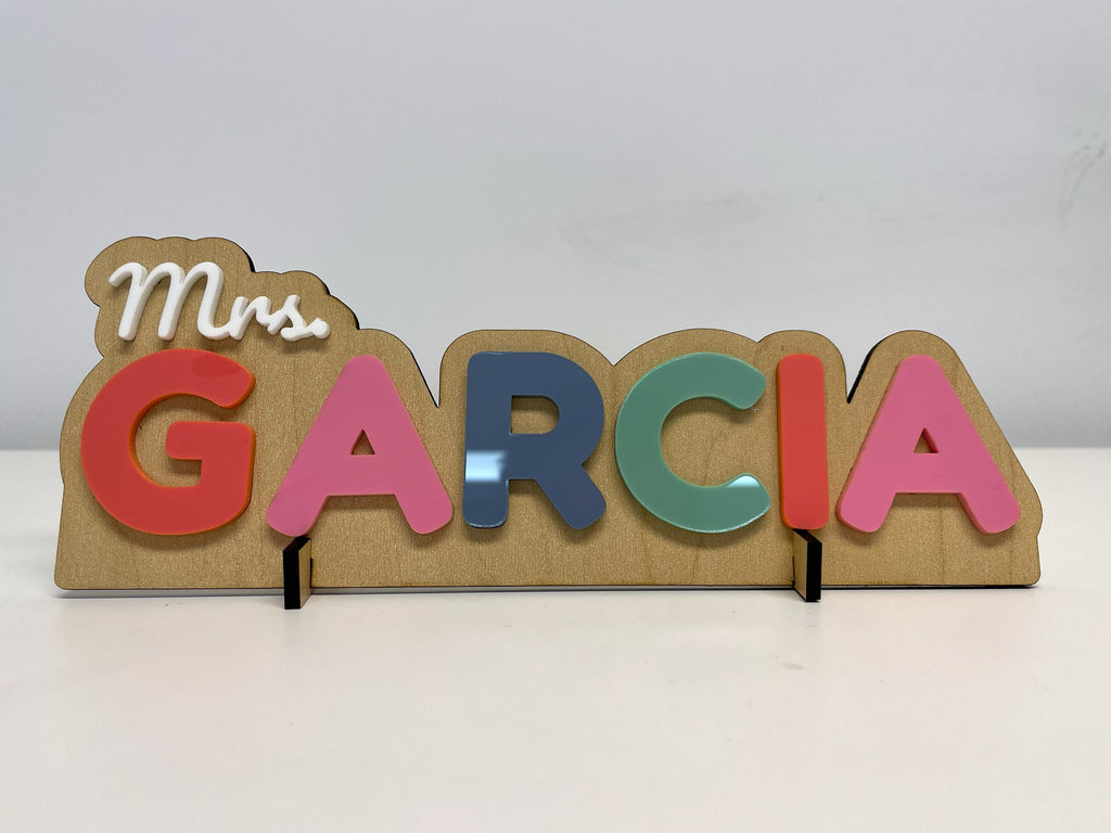 Teacher desk name sign reading Mrs. GARCIA: rainbow raised letters in coral, pink, slate blue and sage under a white script Mrs., on a maple finish base.