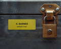 A rectangular yellow nameplate with black text reading S. BARNES and BARNES HVAC, attached to a dark gray metal surface next to a weathered brass padlock with a metal latch mechanism and visible rivets on the mounting plate.