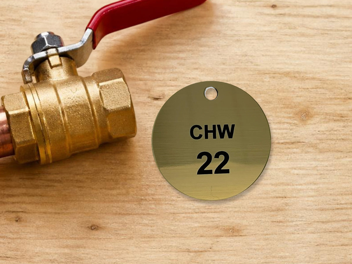 Round brushed gold valve tag engraved CHW 22 in black, lying on a workbench beside a brass ball valve.