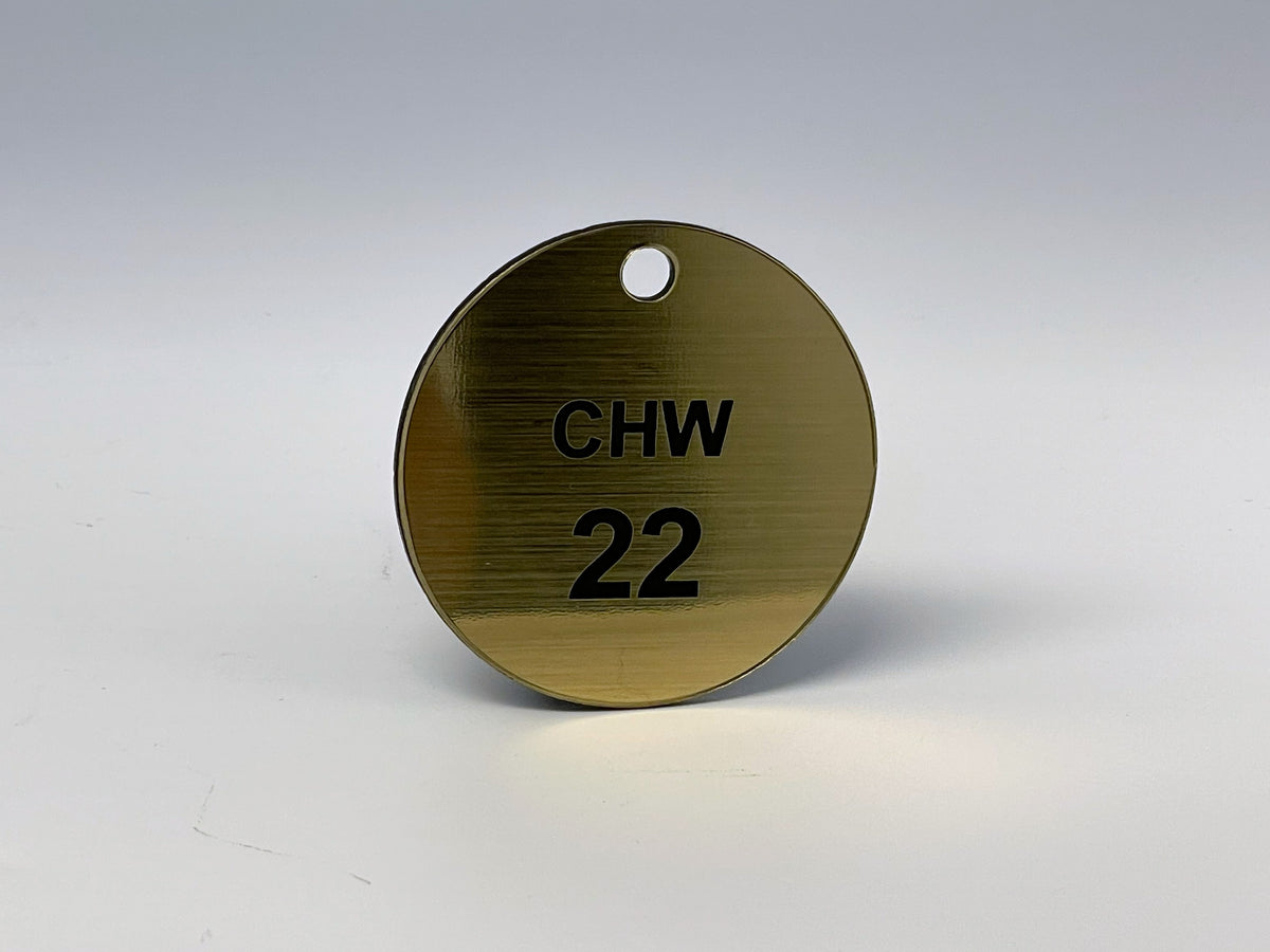 Round brushed gold valve tag engraved CHW 22 in black, with a hole at the top for a chain or S-hook, shown at an angle.