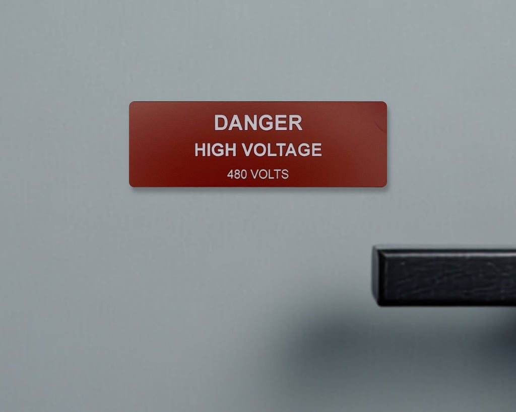 A rectangular red safety sign with white text that reads DANGER HIGH VOLTAGE 480 VOLTS. A black handle is visible on the right.