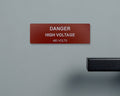 A rectangular red safety sign with white text that reads DANGER HIGH VOLTAGE 480 VOLTS. A black handle is visible on the right.